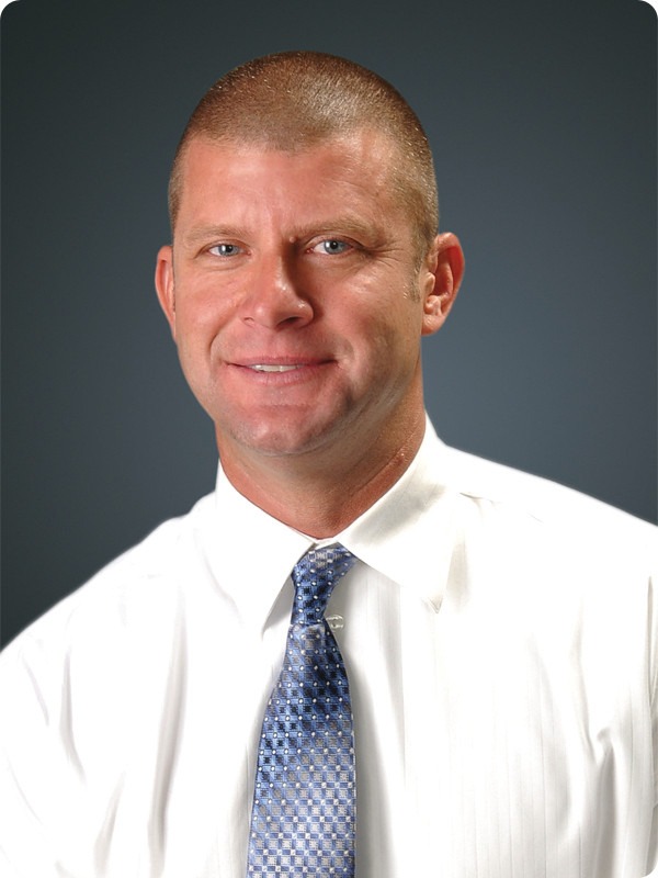 Professional headshot of a man with closely cropped hair wearing a white dress shirt and patterned blue tie, posed against a neutral gray background.